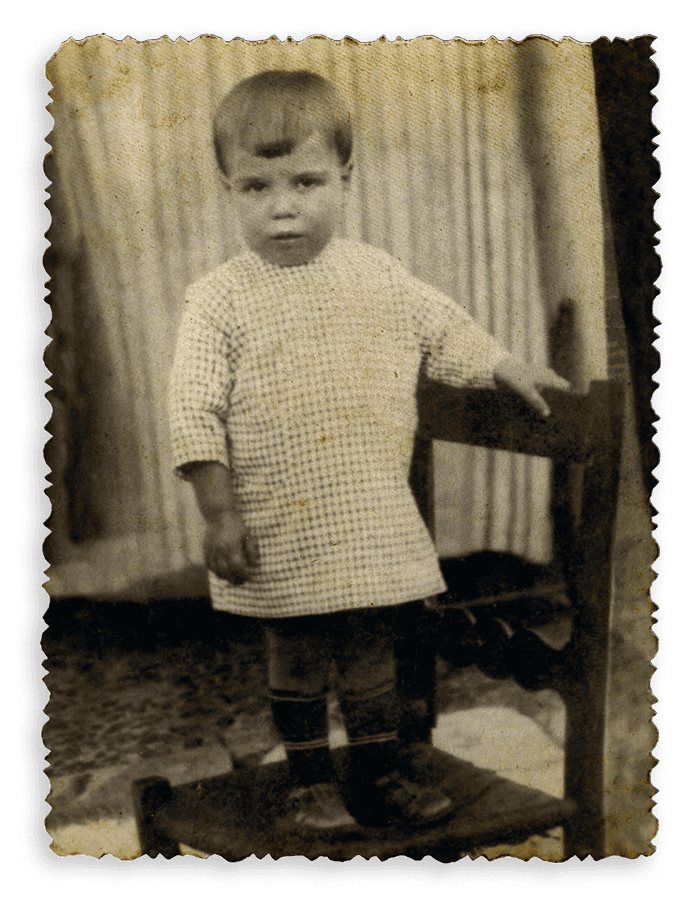 Aged sepia photo of a serious toddler in a checkered shirt standing on a stool next to a chair.