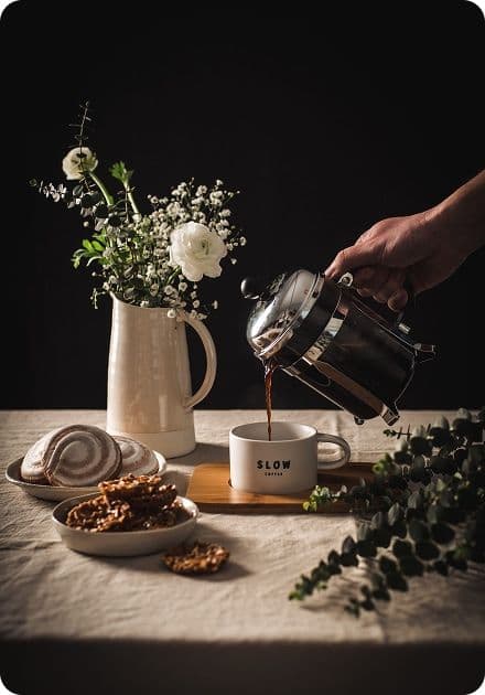 A hand pours coffee from a French press into a white mug labeled "SLOW COFFEE," next to a vase of white flowers and plates of pastries.